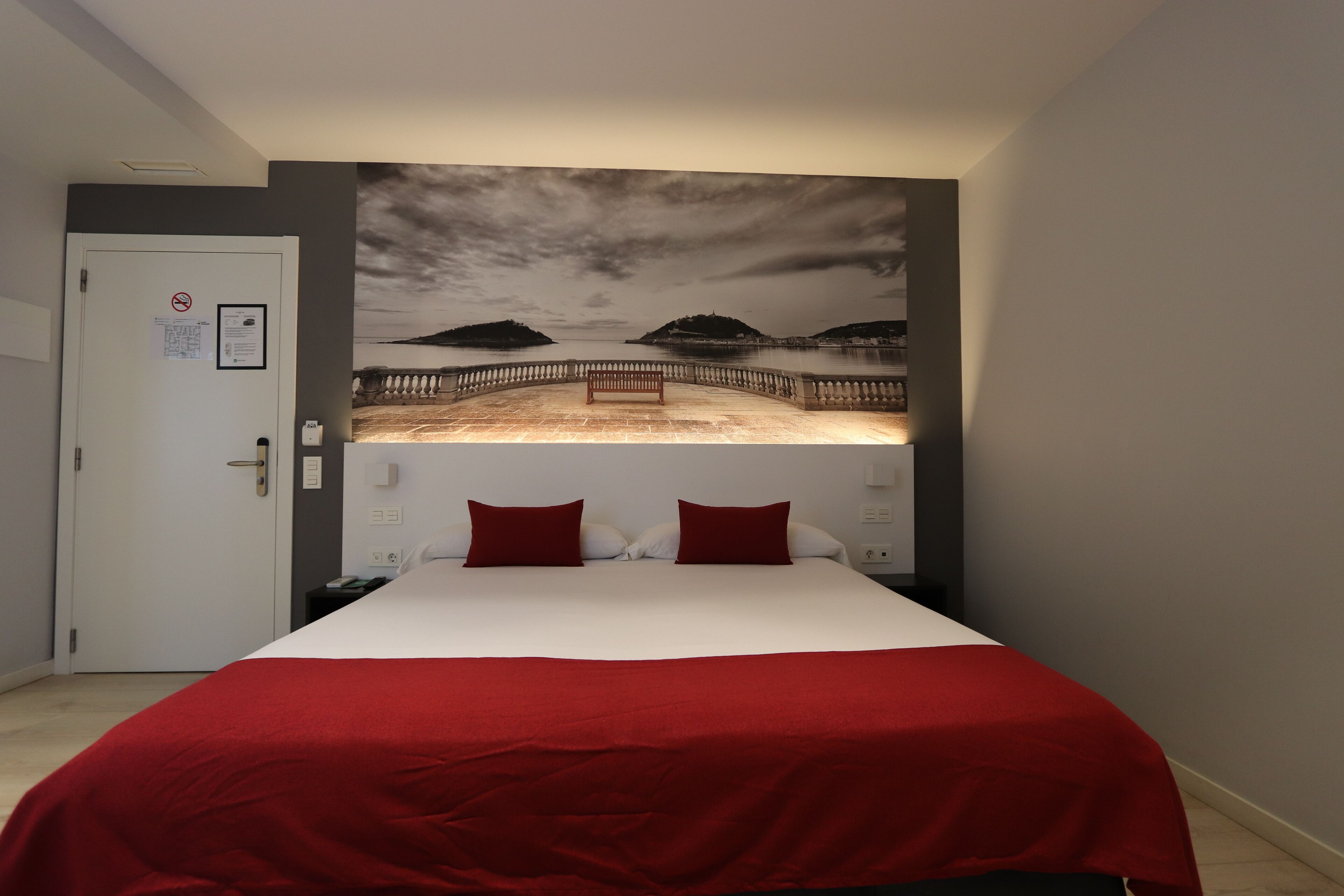 Un dormitorio con una cama grande, una colcha roja, dos almohadas rojas, una mesita de noche, un televisor montado en la pared y una puerta con un cartel de prohibido fumar.
