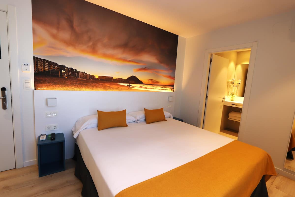 Quarto de hotel com uma cama grande, uma mesa de cabeceira, um quadro na parede e um banheiro visível por uma porta aberta.