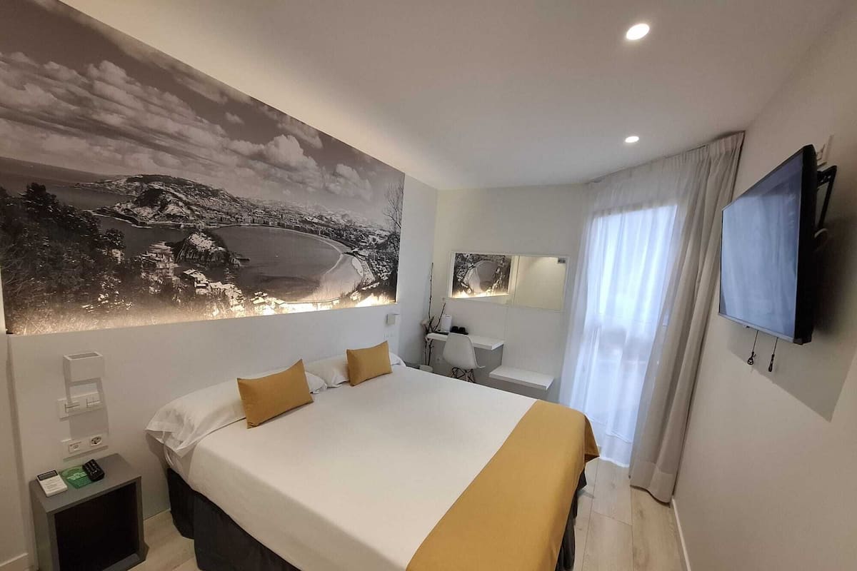 Quarto de hotel moderno com uma cama grande, televisão de tela plana, escrivaninha com cadeira, mesa pequena e uma janela ampla com cortinas.