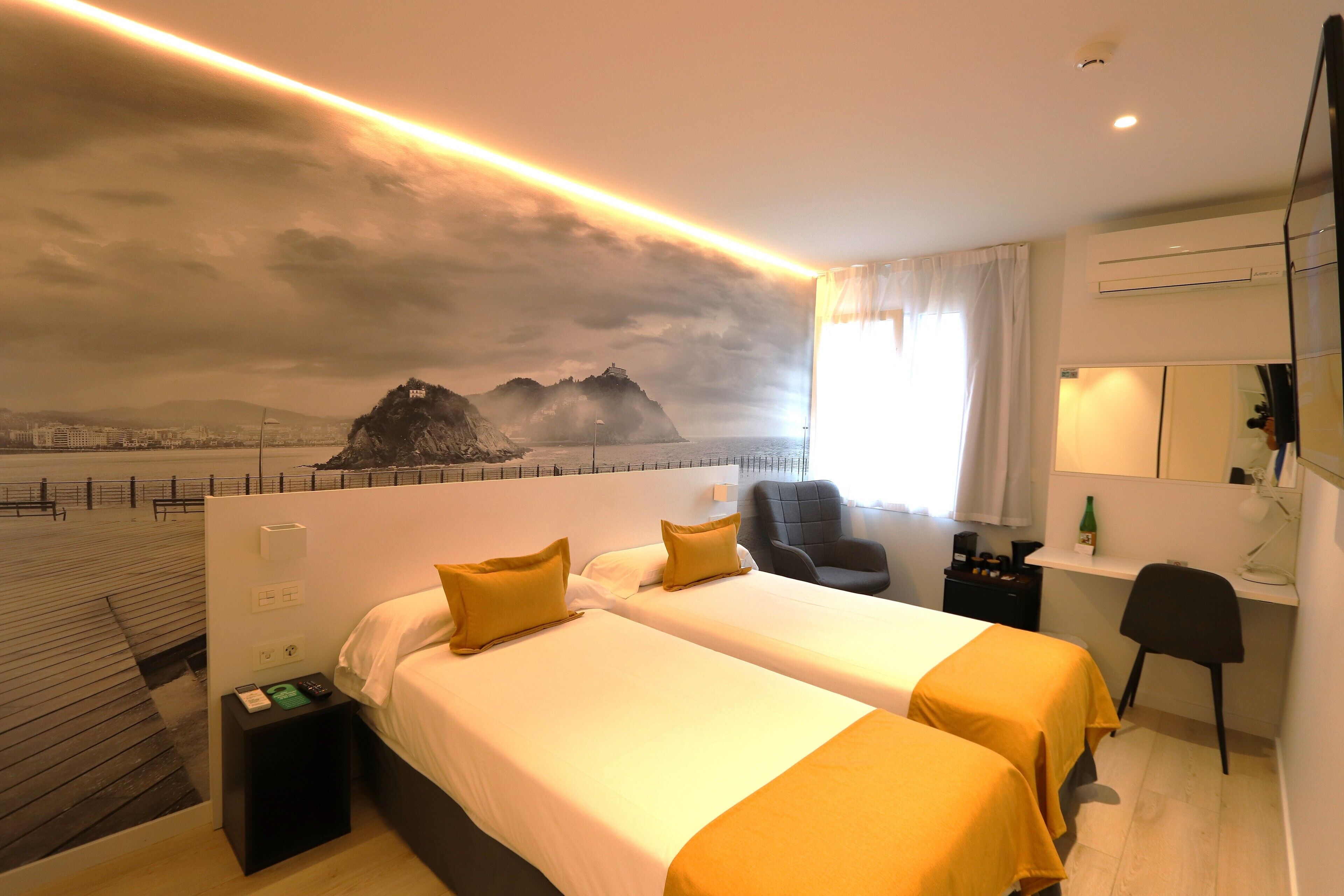 Une chambre d’hôtel avec deux lits, un bureau, une chaise et une vue sur un paysage urbain dominé par des montagnes.