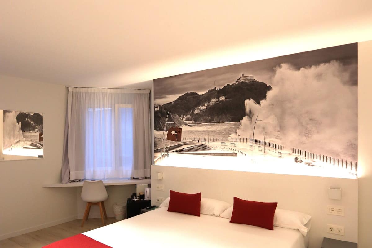 Quarto com cama grande, janela com vista para a costa e escrivaninha com cadeira.