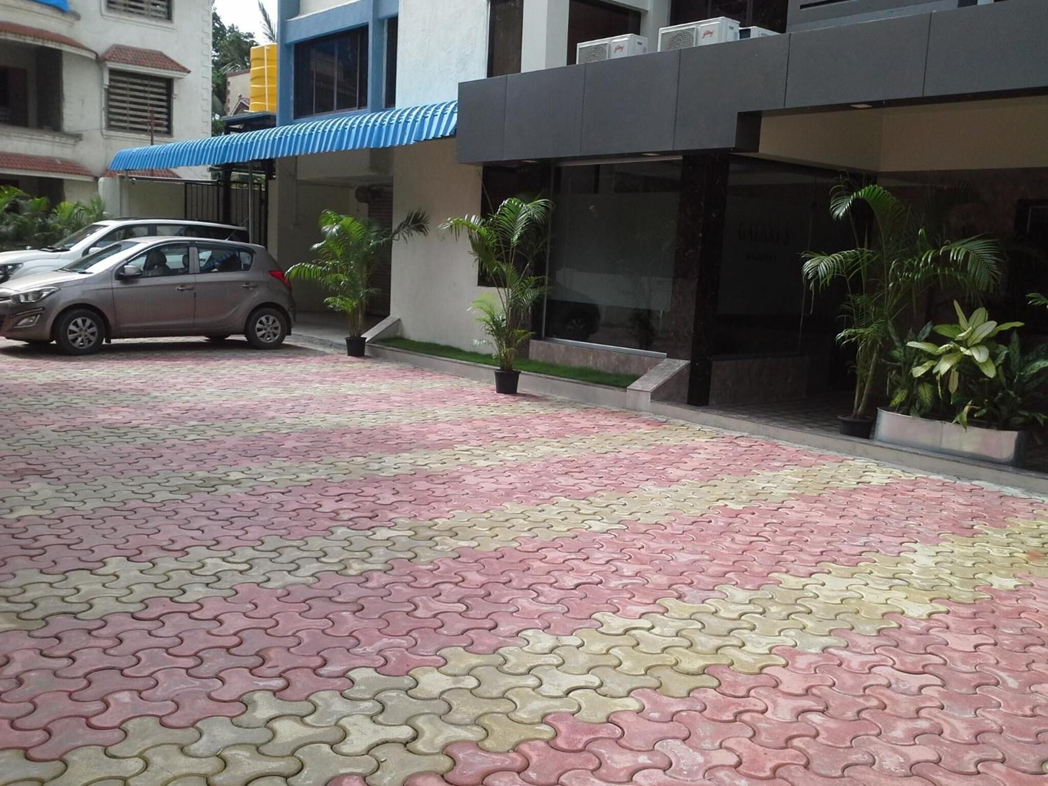 A paved area with interlocking tiles, a parked car, and potted plants near a building with a blue awning.