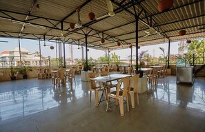 Un café al aire libre con mesas y sillas, techo de metal y plantas en macetas.