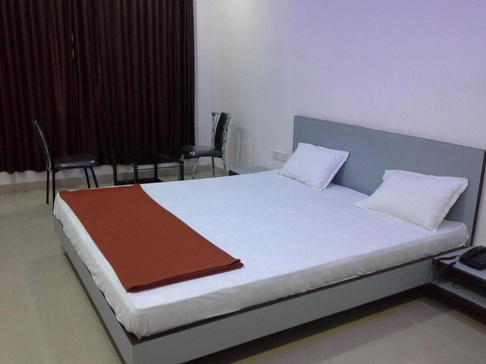 A neatly made bed with white and brown linens, a bedside table with a phone, and a chair in a room with a plain wall and a curtain.