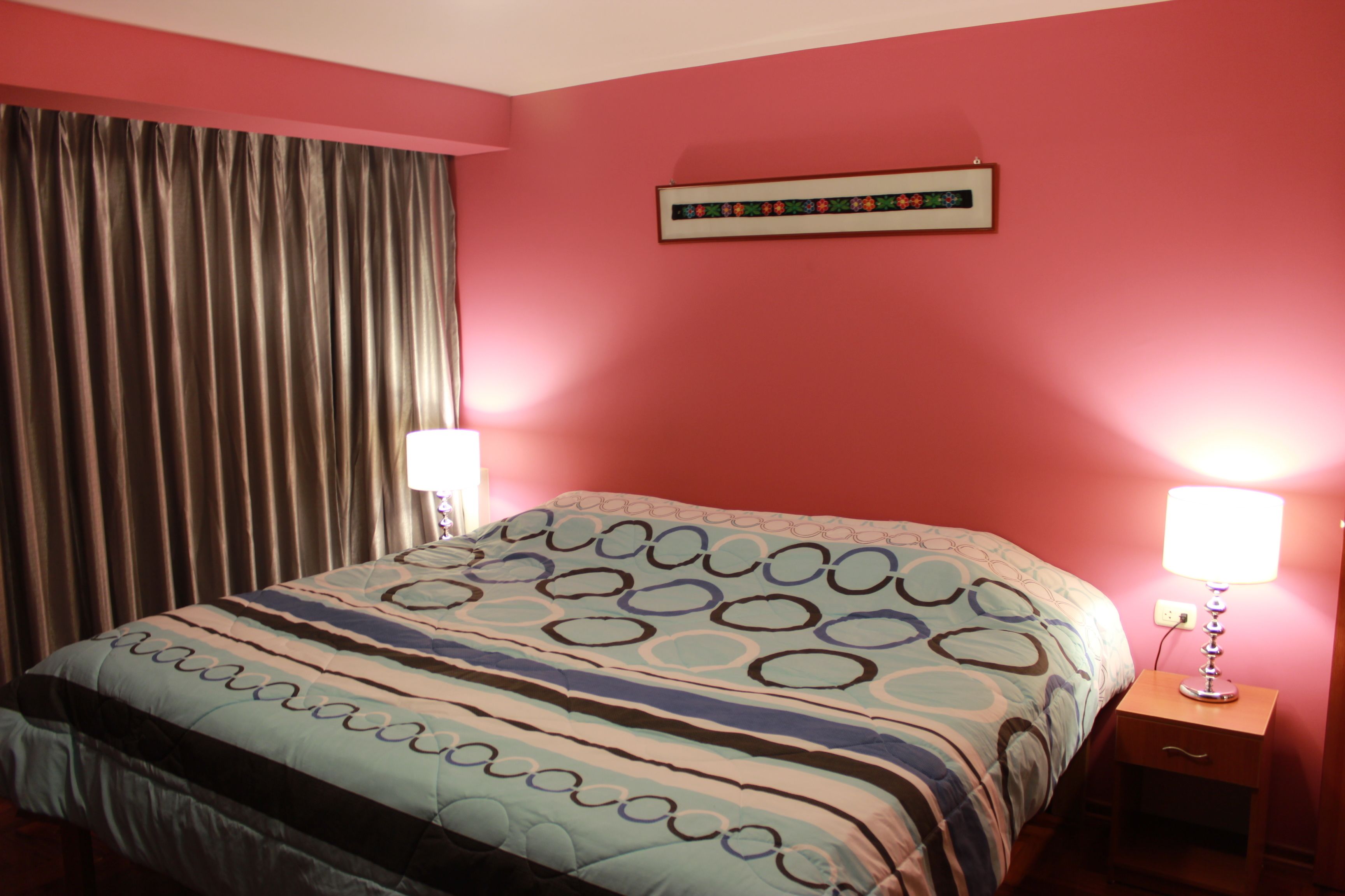 A bedroom with a bed, two bedside lamps, a nightstand, a window with curtains, and a wall-mounted picture.