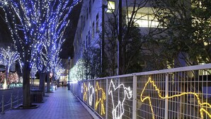 A pathway lined with trees decorated with blue lights, a metal fence with yellow light outlines, and buildings in the background. - APA Hotel Fukushima Ekimae (Fukushima)