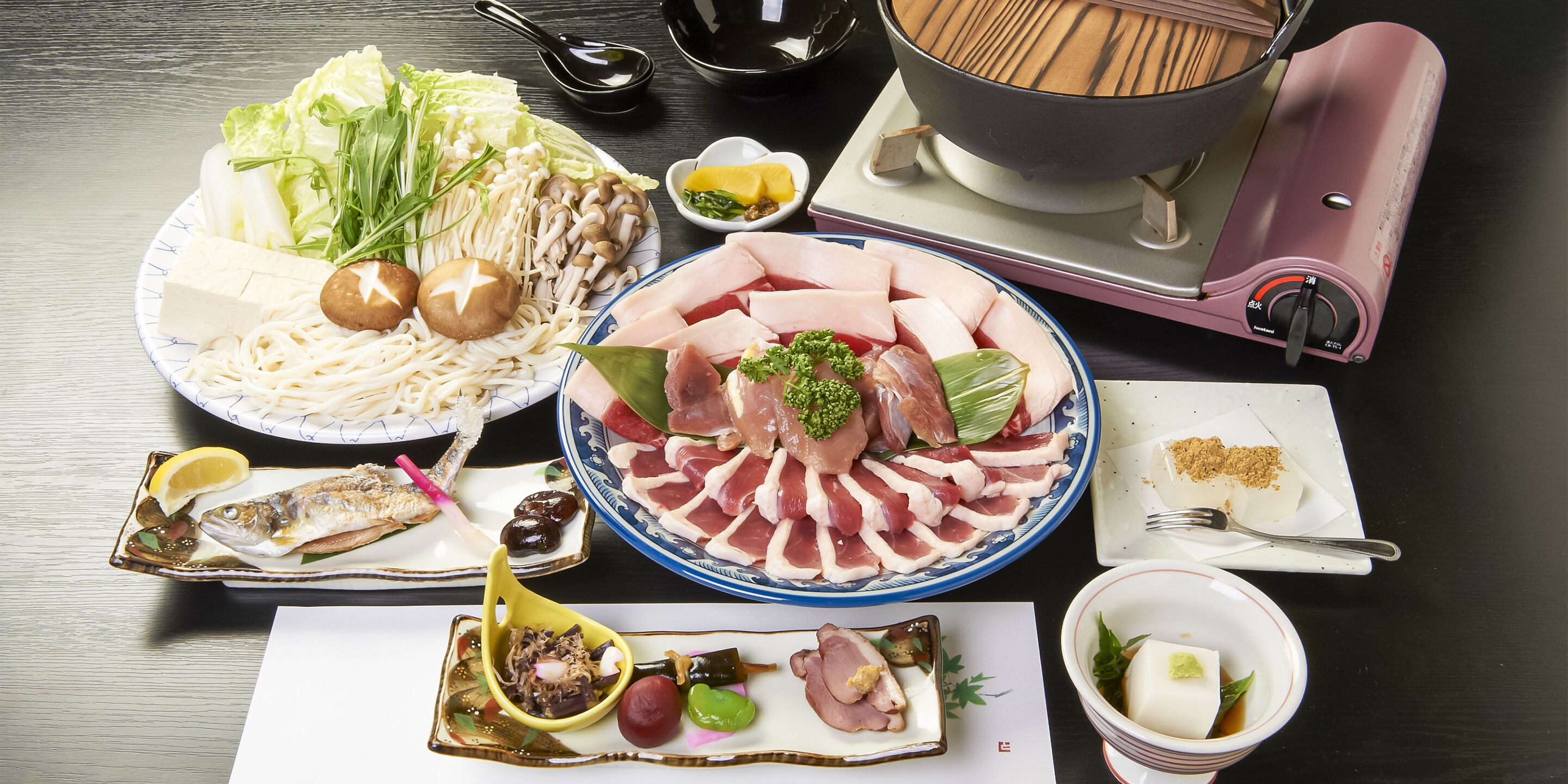 Variedad de platos que incluyen fideos, sashimi y mariscos, dispuestos sobre una mesa con un recipiente caliente y palillos.