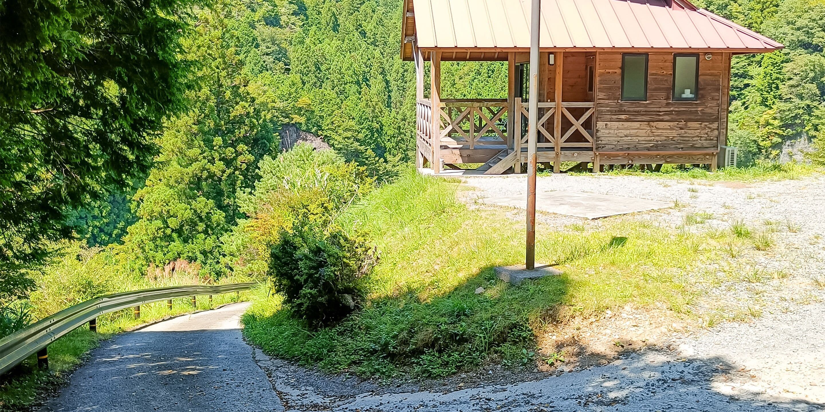 A wooden cabin with a metal roof, surrounded by lush greenery and a gravel path leading to it.