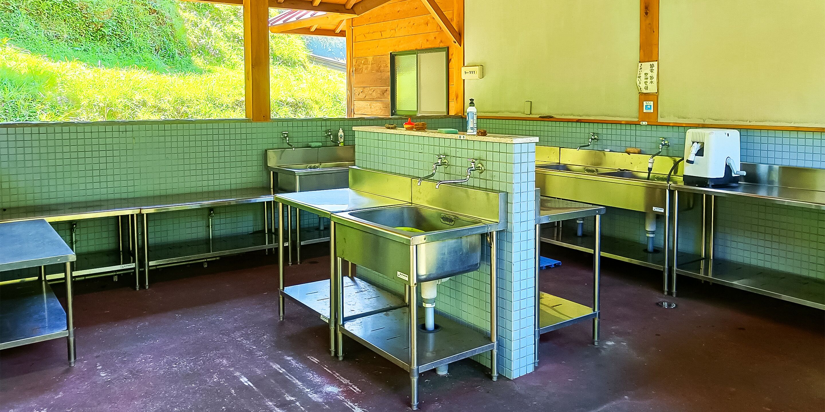 Una cocina con varios lavamanos de acero inoxidable, una cafetera y una ventana con vistas a zonas verdes.