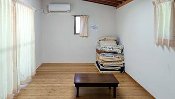 A room with a wooden floor, a small table, a window with curtains, an air conditioning unit, and a stack of folded clothes against the wall.