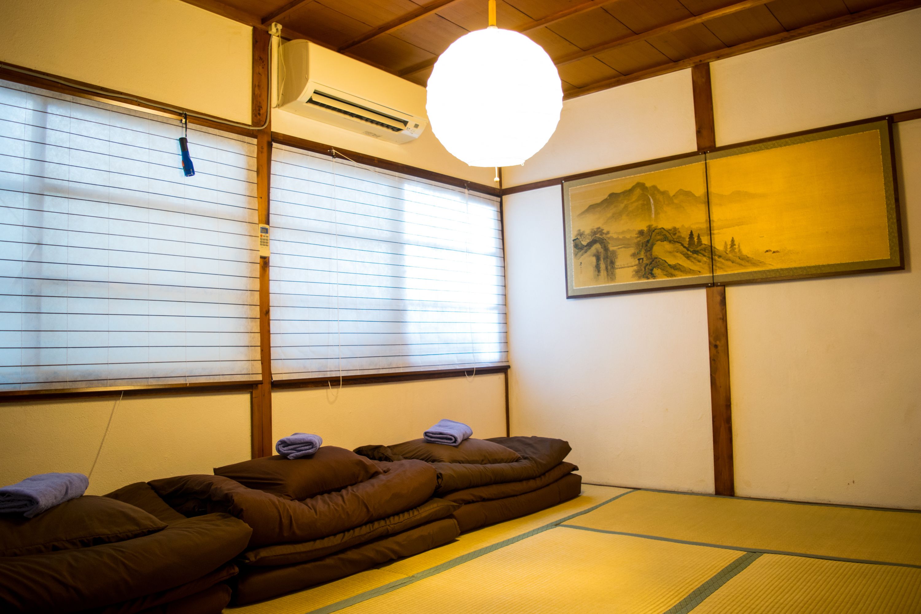 A traditional Japanese room with tatami flooring, a futon bed, a hanging light fixture, and a painting on the wall.