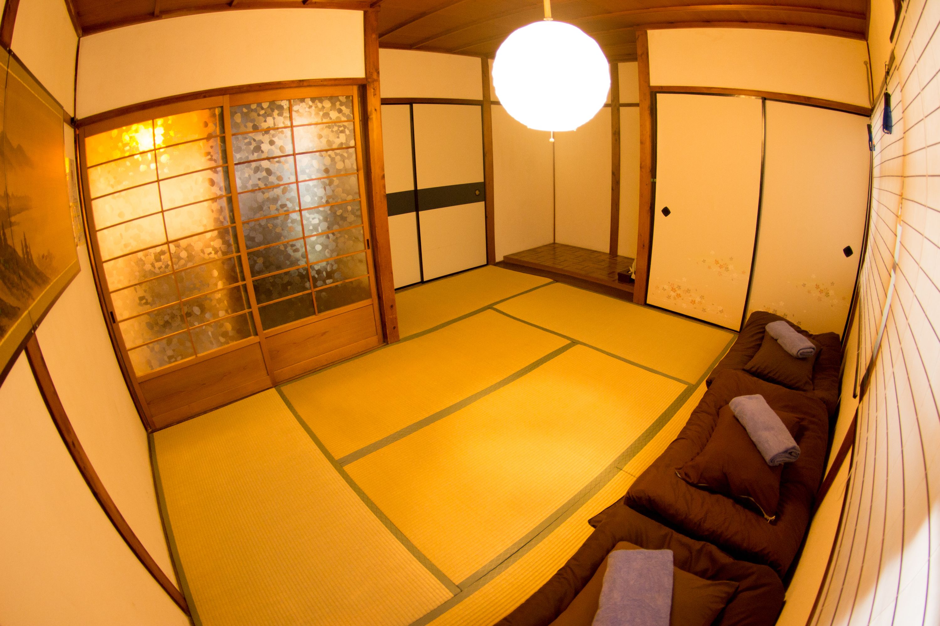 Une pièce avec un sol recouvert de tatamis, un futon servant de lit, des portes coulissantes et un luminaire suspendu.
