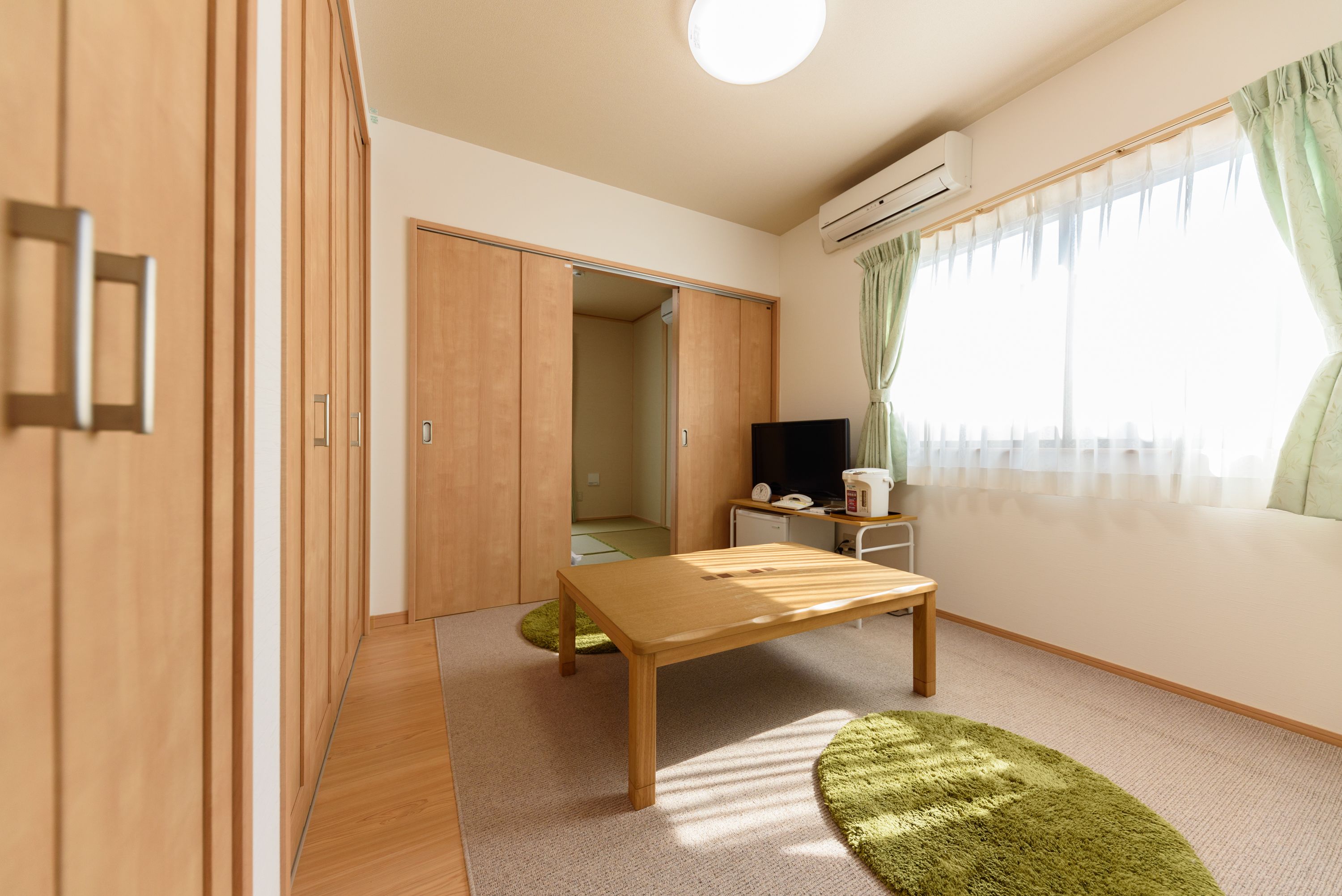 Ein Zimmer mit Holzschrank, kleinem Tisch, grünem Teppich und einem Fenster mit Vorhängen.