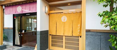 A traditional Japanese building with a wooden entrance, stone flooring, and a green plant on the right.