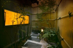 A narrow pathway with stepping stones, surrounded by bamboo and ferns, leading to a lit window. - Hosta Nijo Sen (Kyoto)