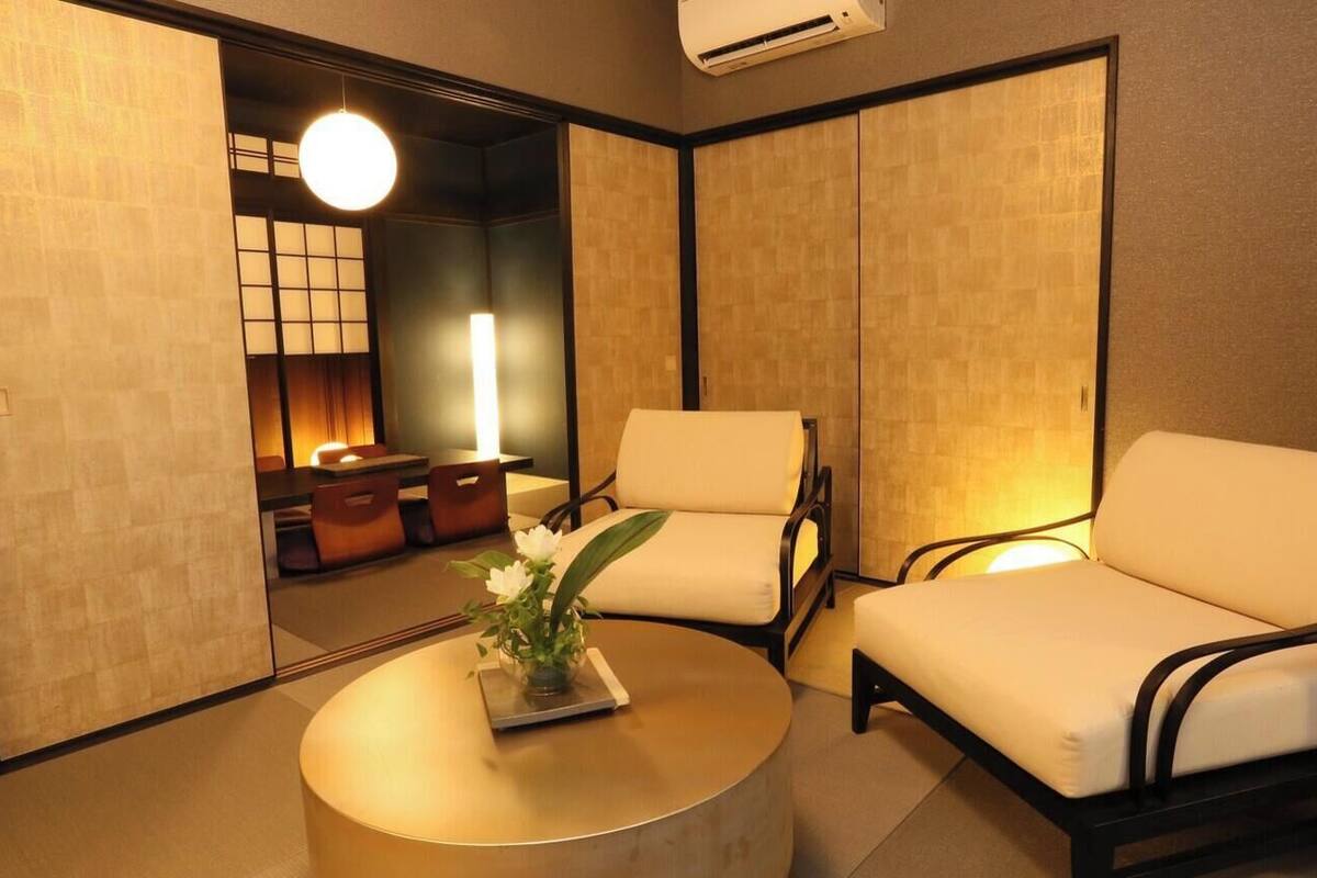 A room with two chairs, a round table with a vase of flowers, and a view through a shoji screen into a dining area.
