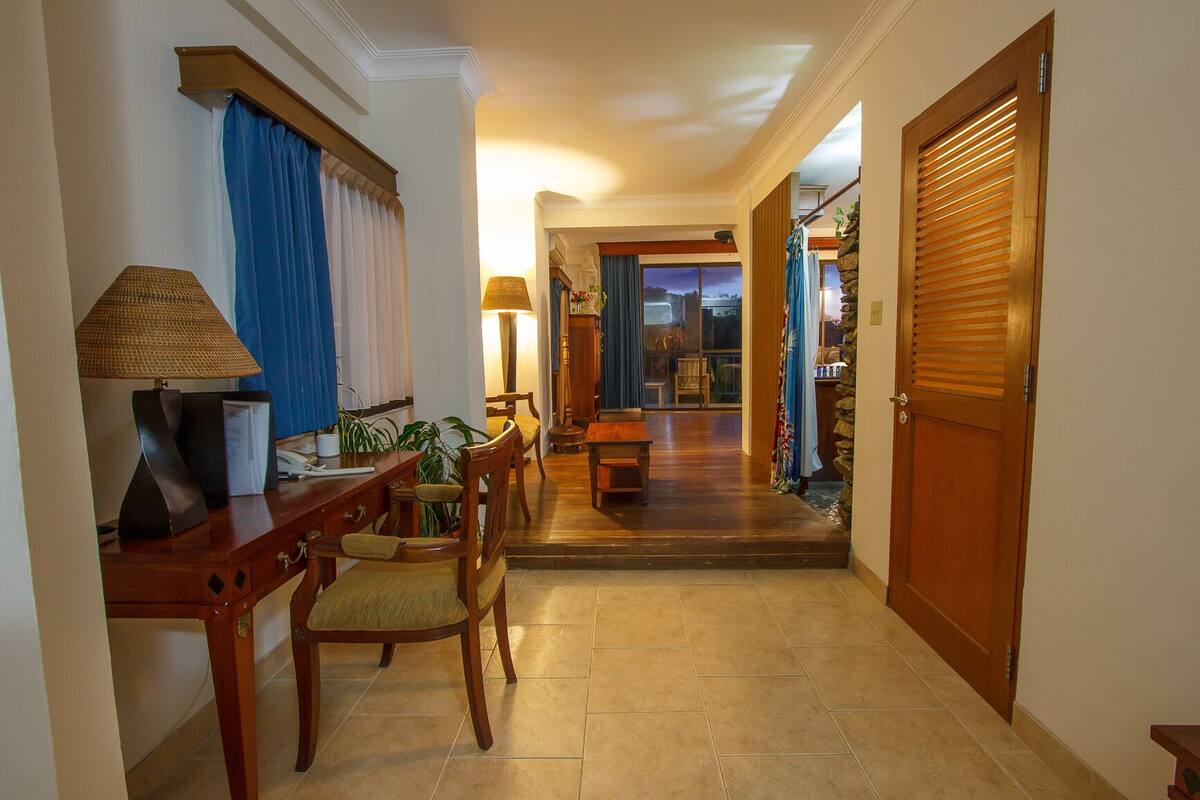 A hotel lobby with a wooden desk, chairs, a lamp, and a view of a room through a glass door.