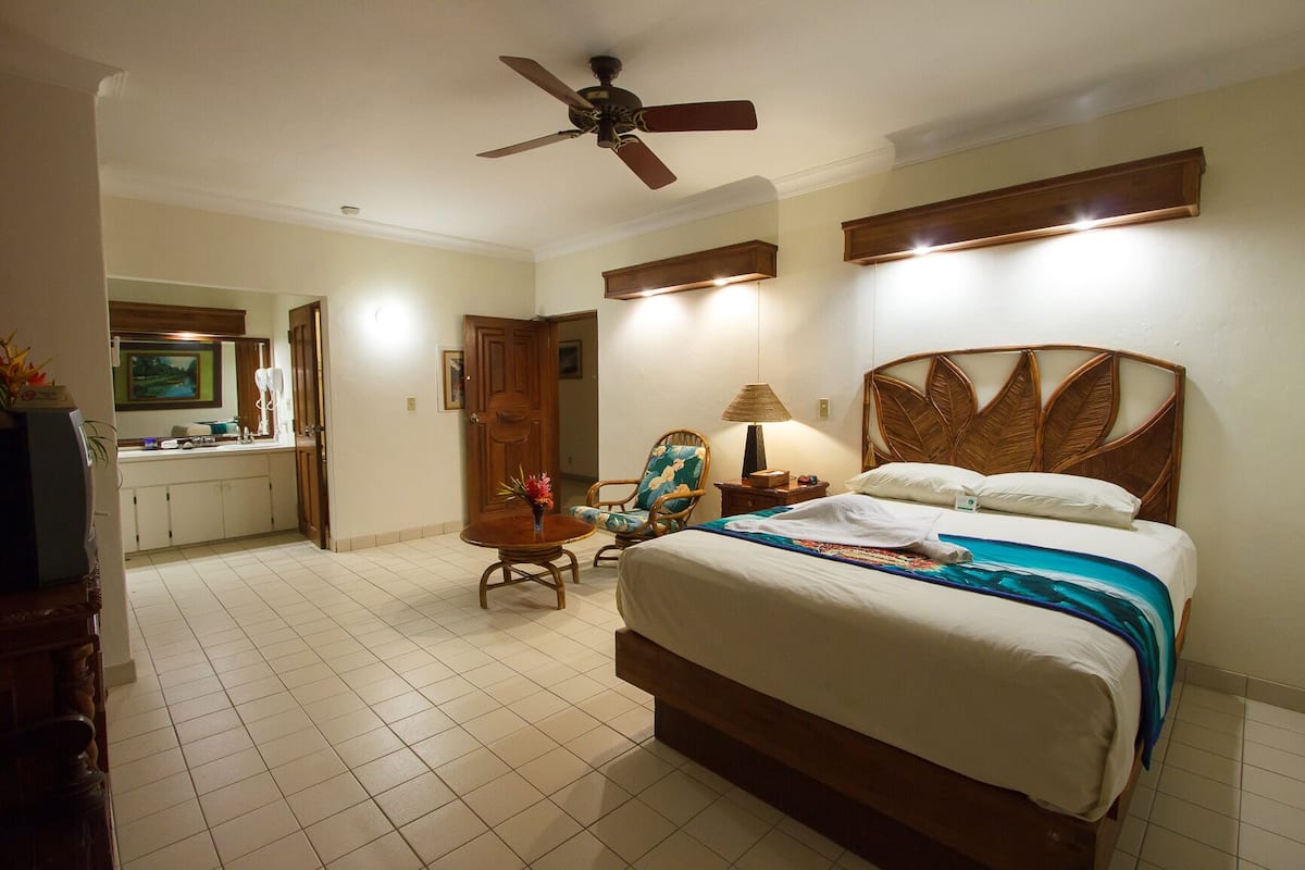 A hotel room with a bed, a chair, a desk, a ceiling fan, and a bathroom visible through an open door.