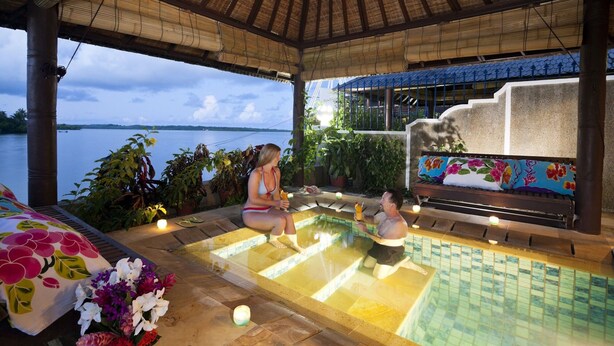 Un espace piscine avec vue sur l’eau, un coin salon abrité et un banc agrémenté d’un coussin à motifs floraux.