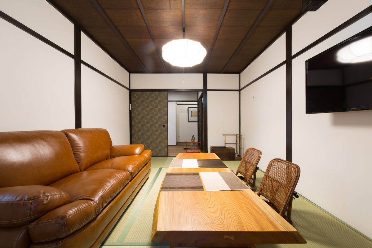 A traditional Japanese-style room with tatami flooring, a wooden table, a brown leather sofa, and a television mounted on the wall.