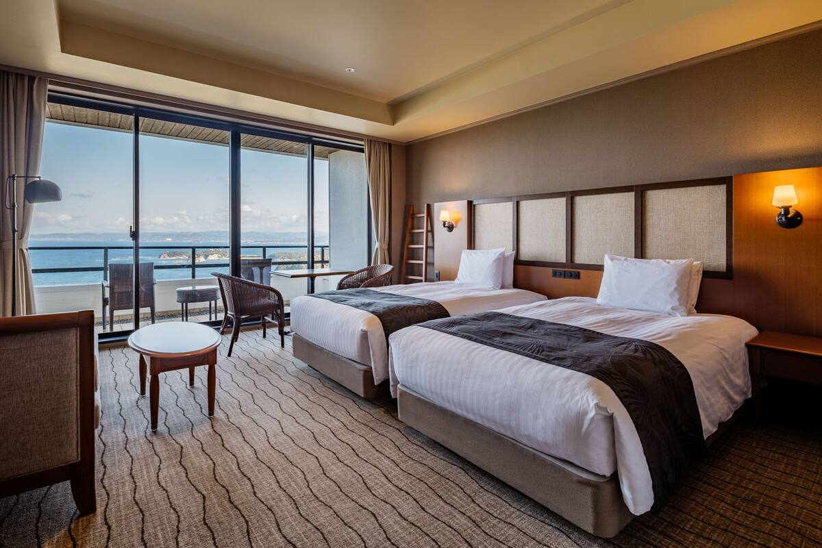 A hotel room with two beds, a small table, a chair, a view of the ocean, and a balcony.