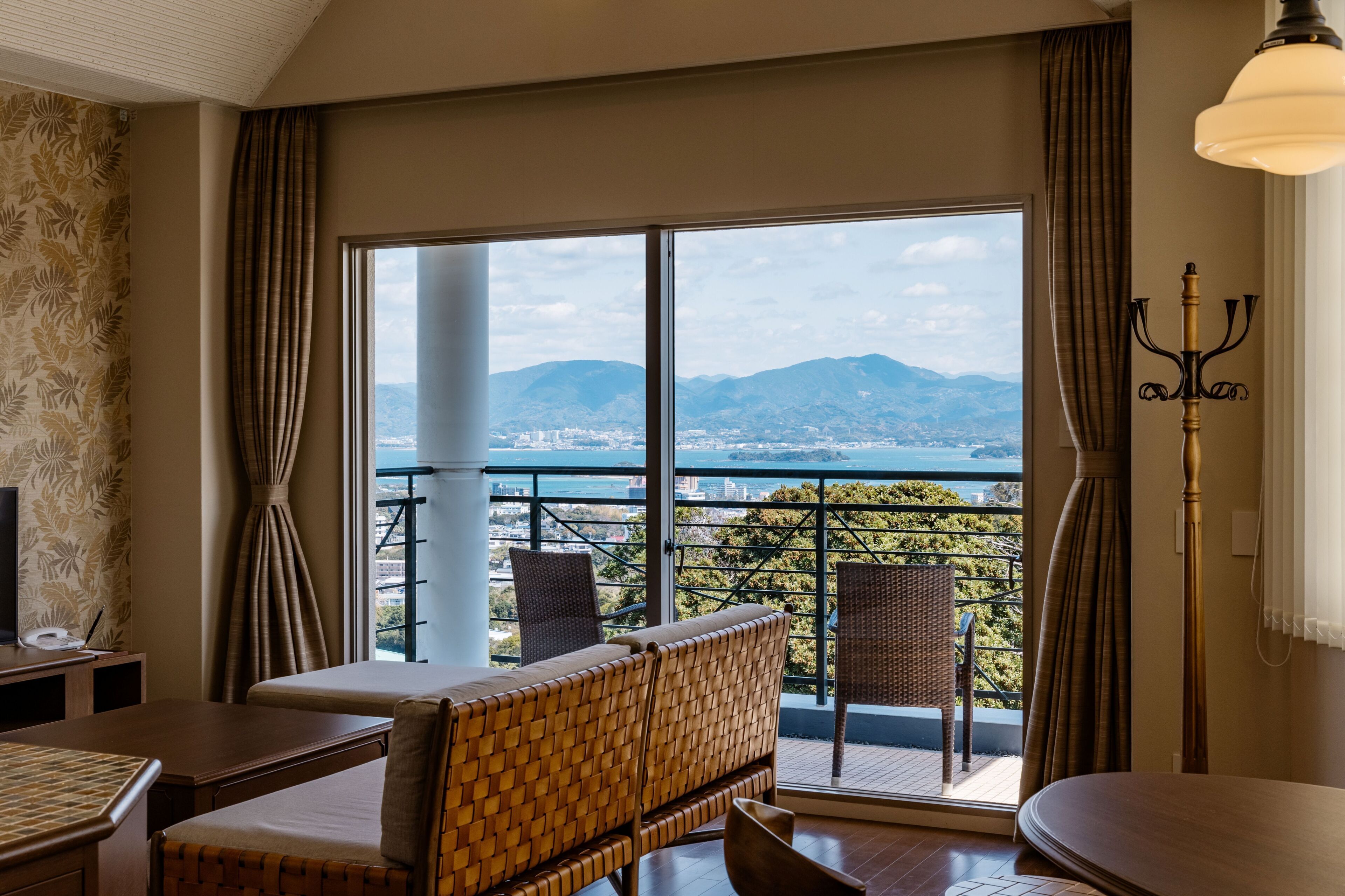 A hotel room with a balcony, wicker furniture, a table, and a view of mountains.