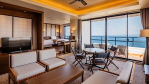 A hotel room with a TV, sofa, dining table, chairs, and a balcony with a view of the sea.