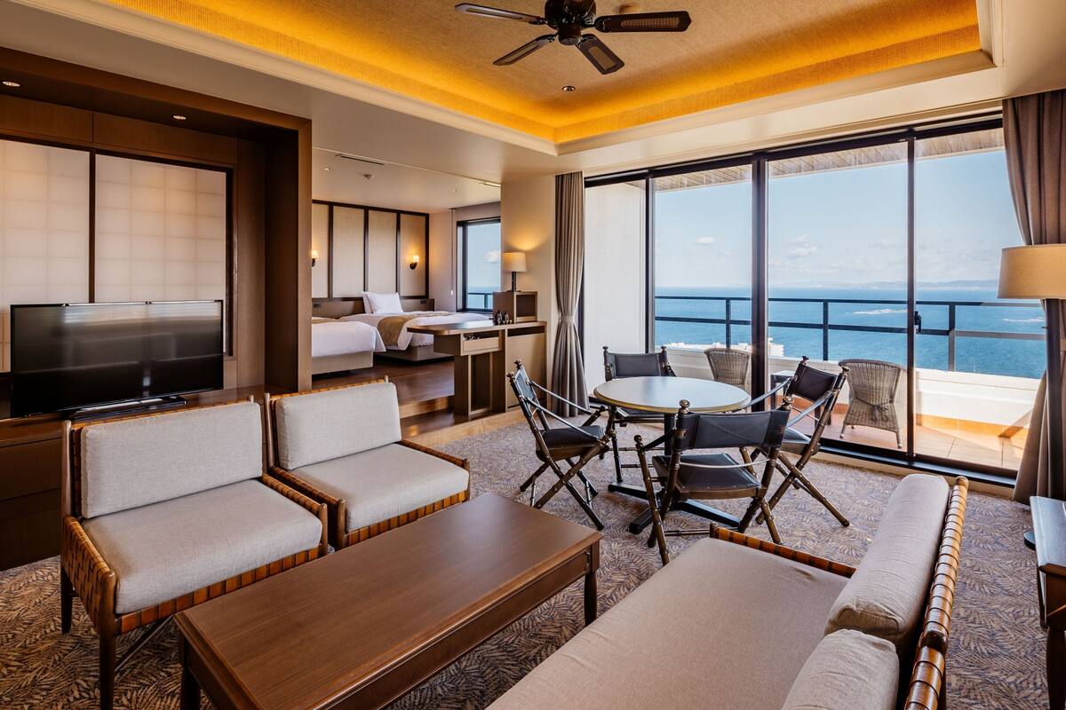 A hotel room with a TV, sofa, dining table, chairs, and a balcony with a view of the sea.