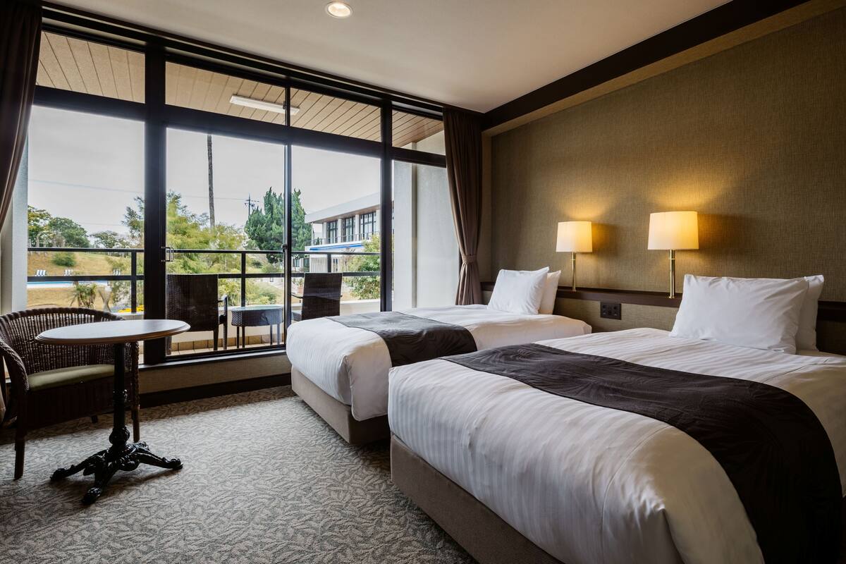 A hotel room with two beds, a small table, and a window with a view of buildings and trees.