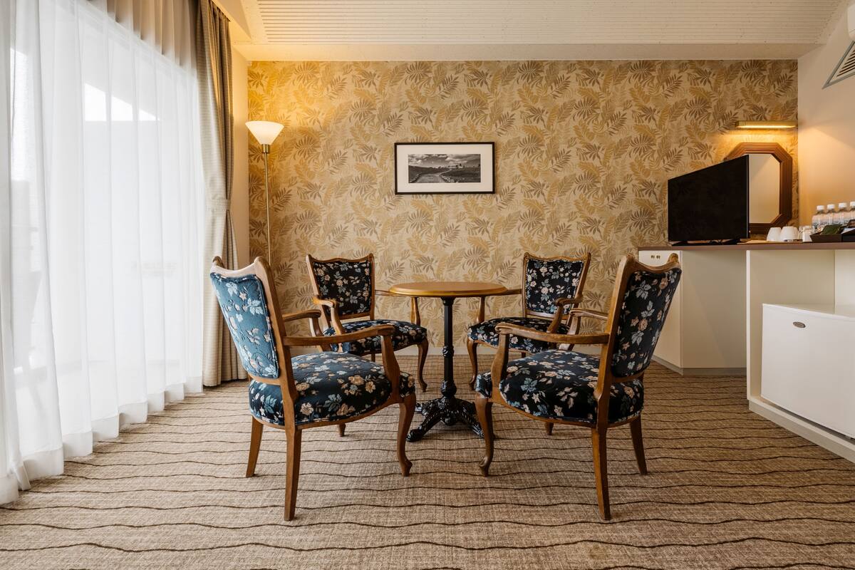 A hotel room with a dining table, four chairs, a television, a framed picture on the wall, and a lamp.