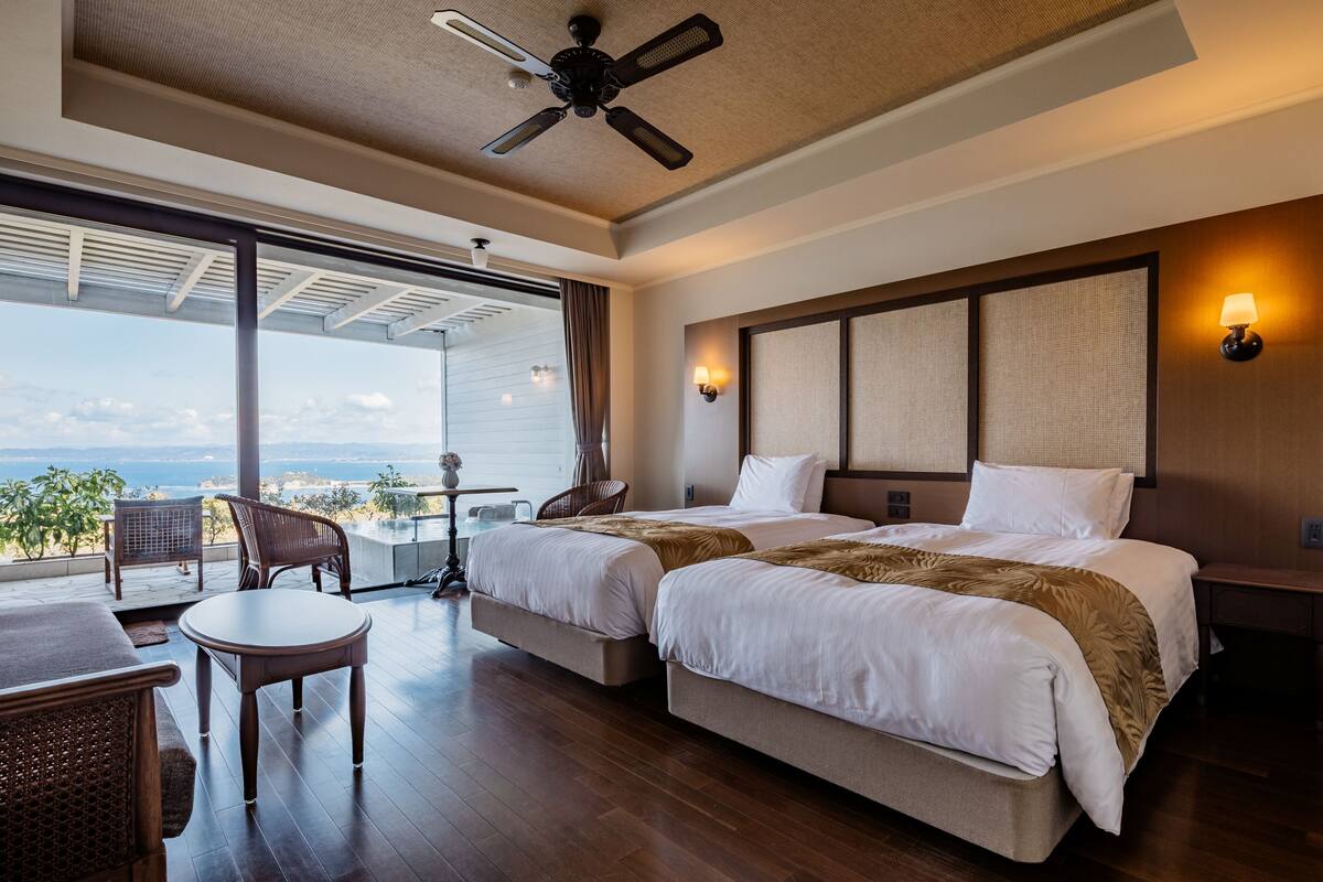 A hotel room with two beds, a ceiling fan, a balcony with a view, and a small table with chairs.