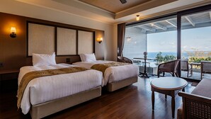 A hotel room with two beds, a ceiling fan, a balcony with a table and chairs, and a view of the ocean.