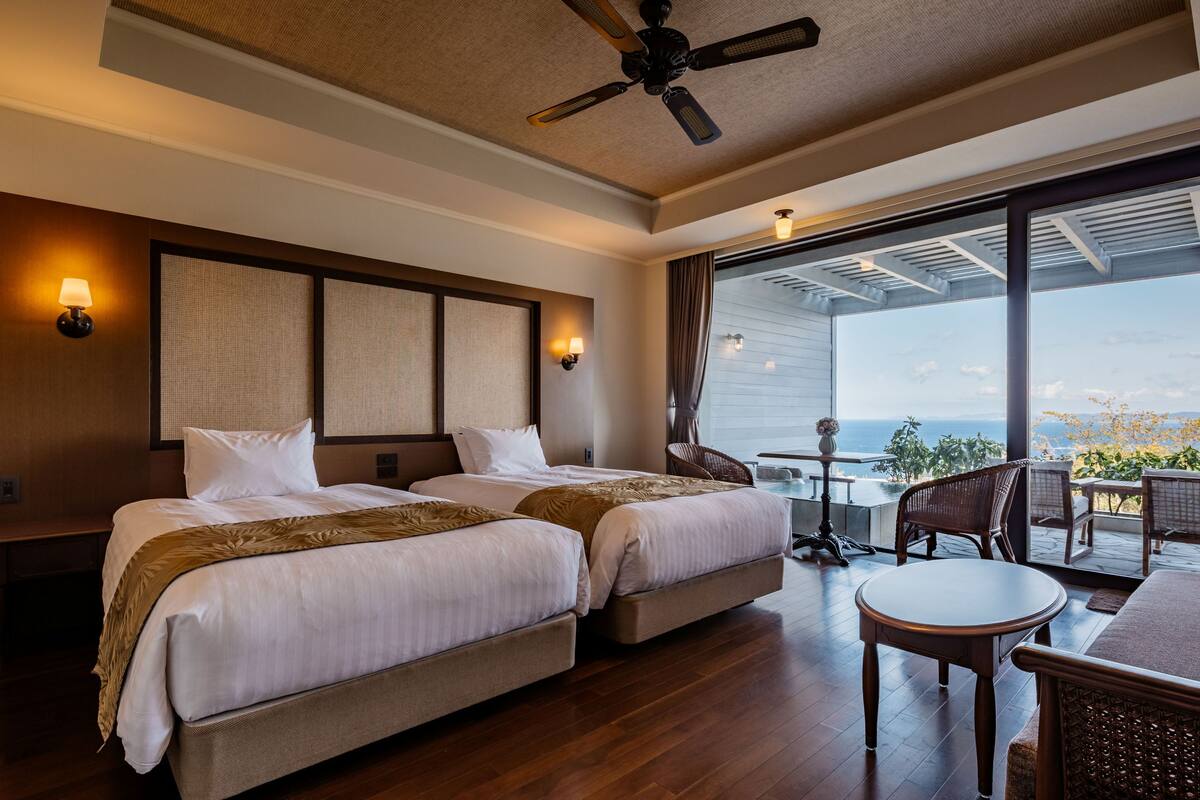 A hotel room with two beds, a ceiling fan, a balcony with a table and chairs, and a view of the ocean.