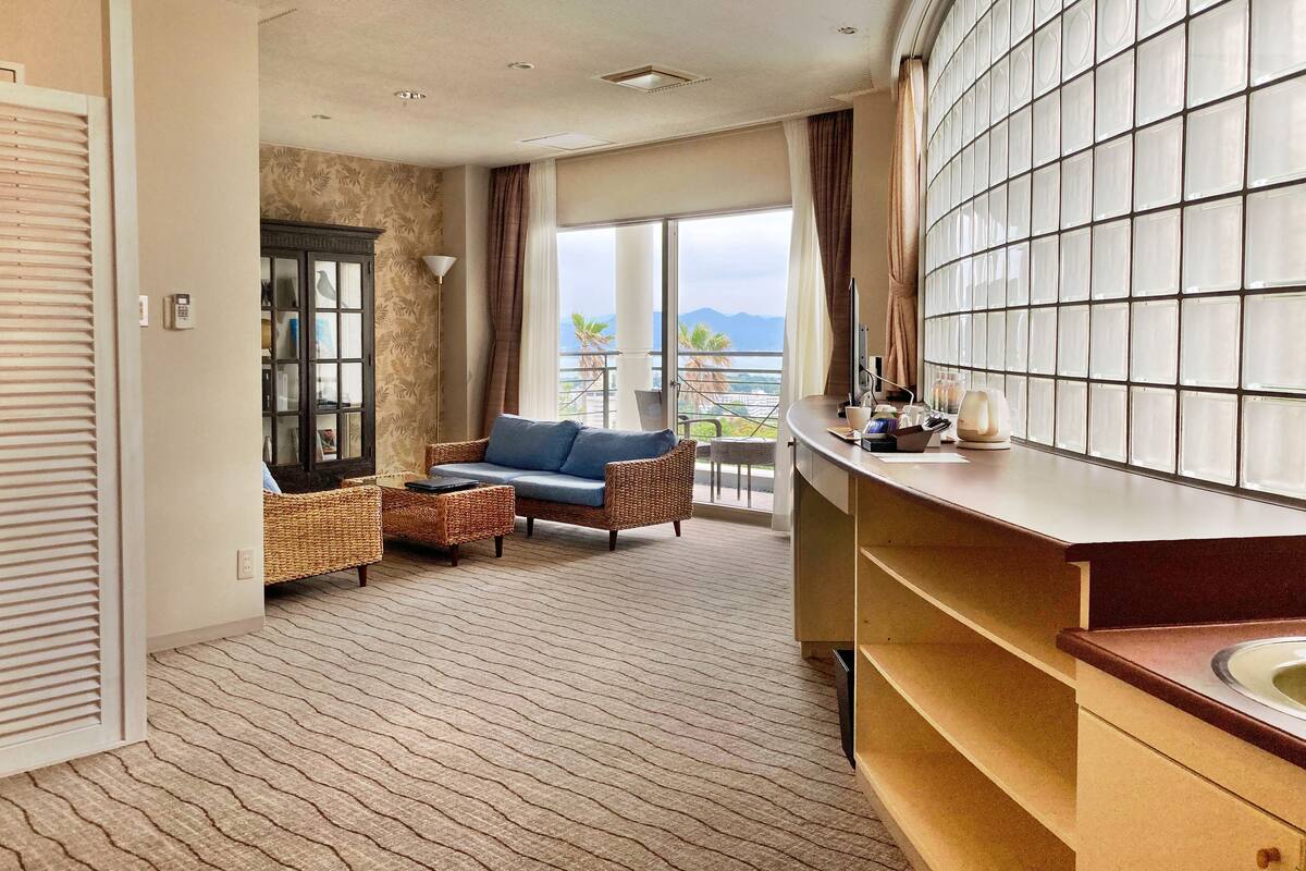 A hotel room with a balcony, a sofa, a wicker chair, a large mirror, and a view of the mountains.