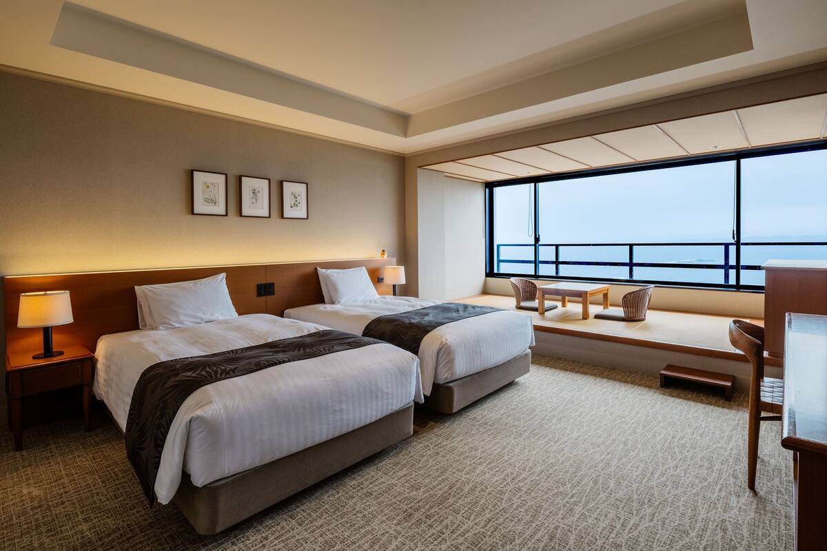 A hotel room with two beds, a desk, a chair, a window with a view, and artwork on the wall.