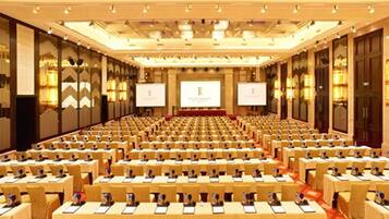 Un amplio salón de conferencias con filas de mesas y sillas, equipado con pantallas de presentación y un escenario.