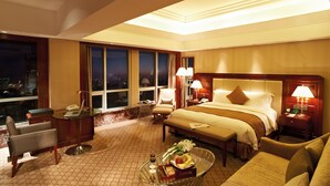 Habitación de hotel con una cama grande, un escritorio con una computadora portátil, una silla, un sofá y una mesa de comedor con un frutero y vino.