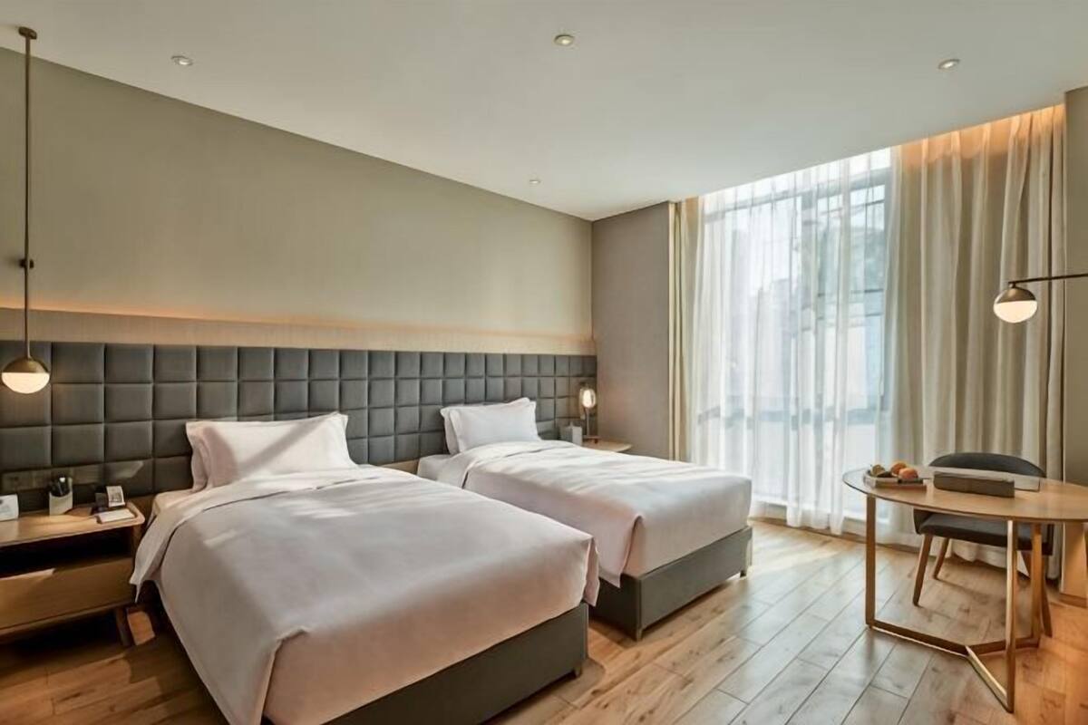 Habitación de hotel con dos camas, suelo de madera, un gran ventanal con cortinas, una mesita de noche y una pequeña mesa redonda.