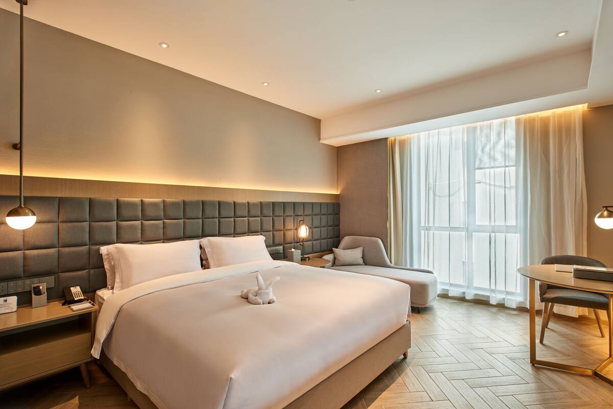 Una habitación de hotel moderna con una cama grande, una mesita de noche con lámpara, un escritorio pequeño y una silla.