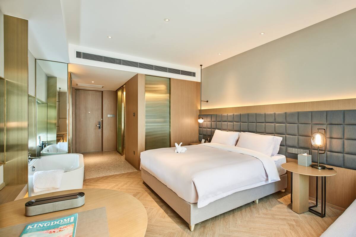 Una habitación de hotel moderna con una cama grande, una mesita de noche con lámpara, una mesa redonda y un espejo.