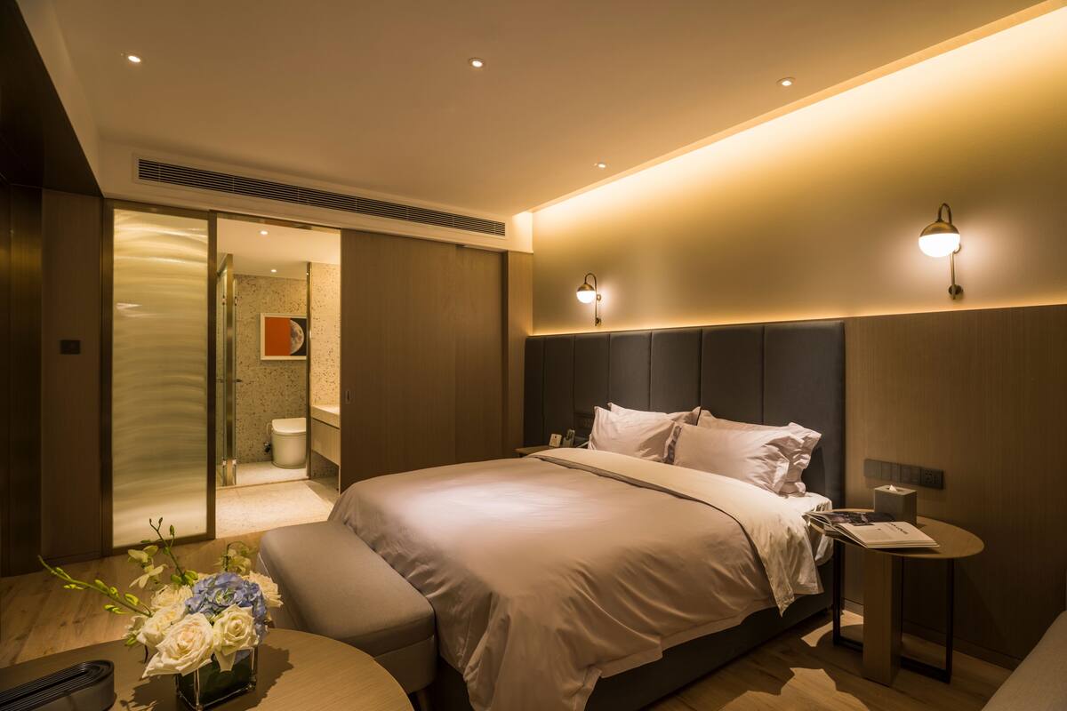 Una habitación de hotel moderna con una cama grande, mesitas de noche, una pequeña zona de estar y un baño visible a través de una puerta de cristal.