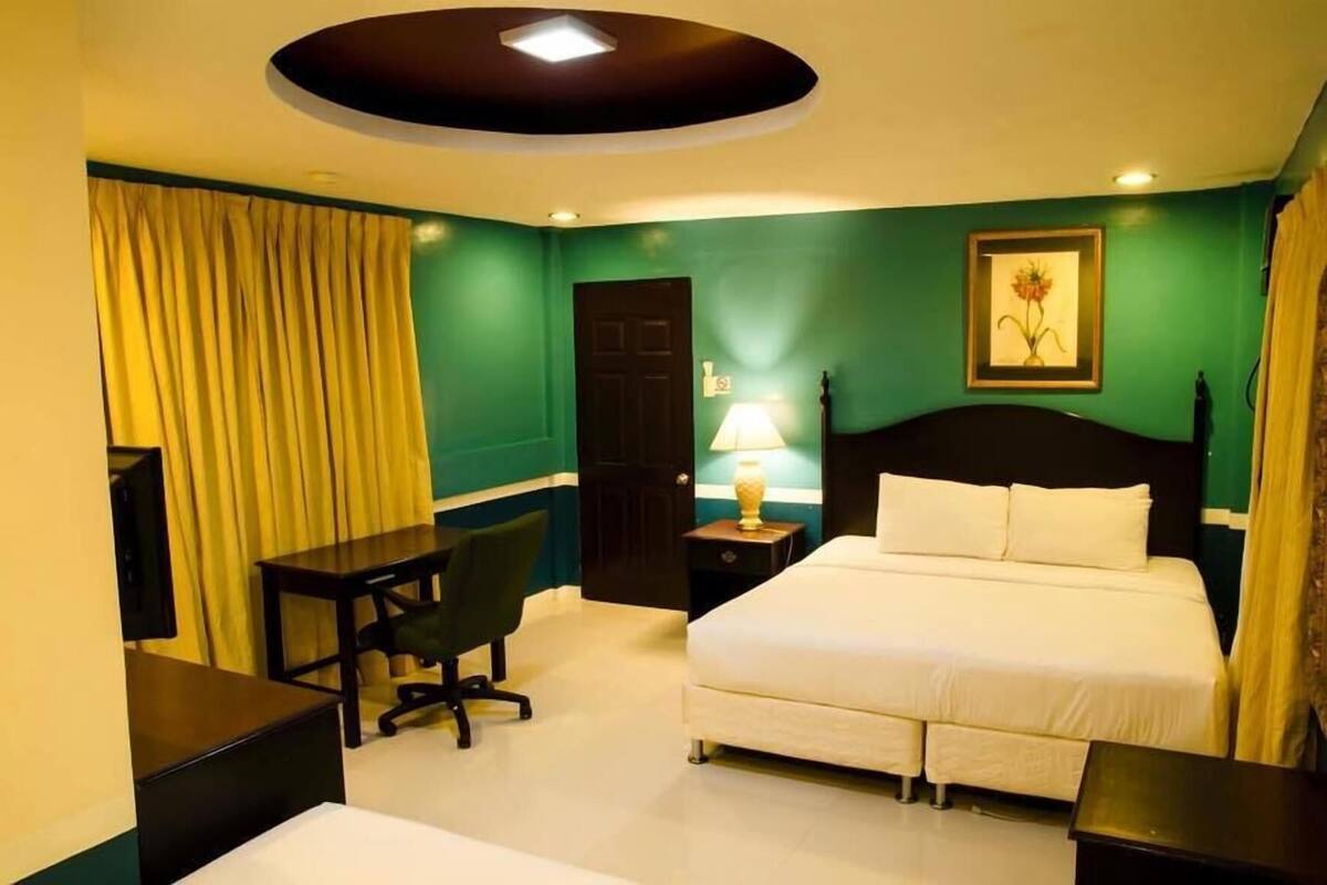 A hotel room with a bed, a desk, a chair, a nightstand with a lamp, and a door.