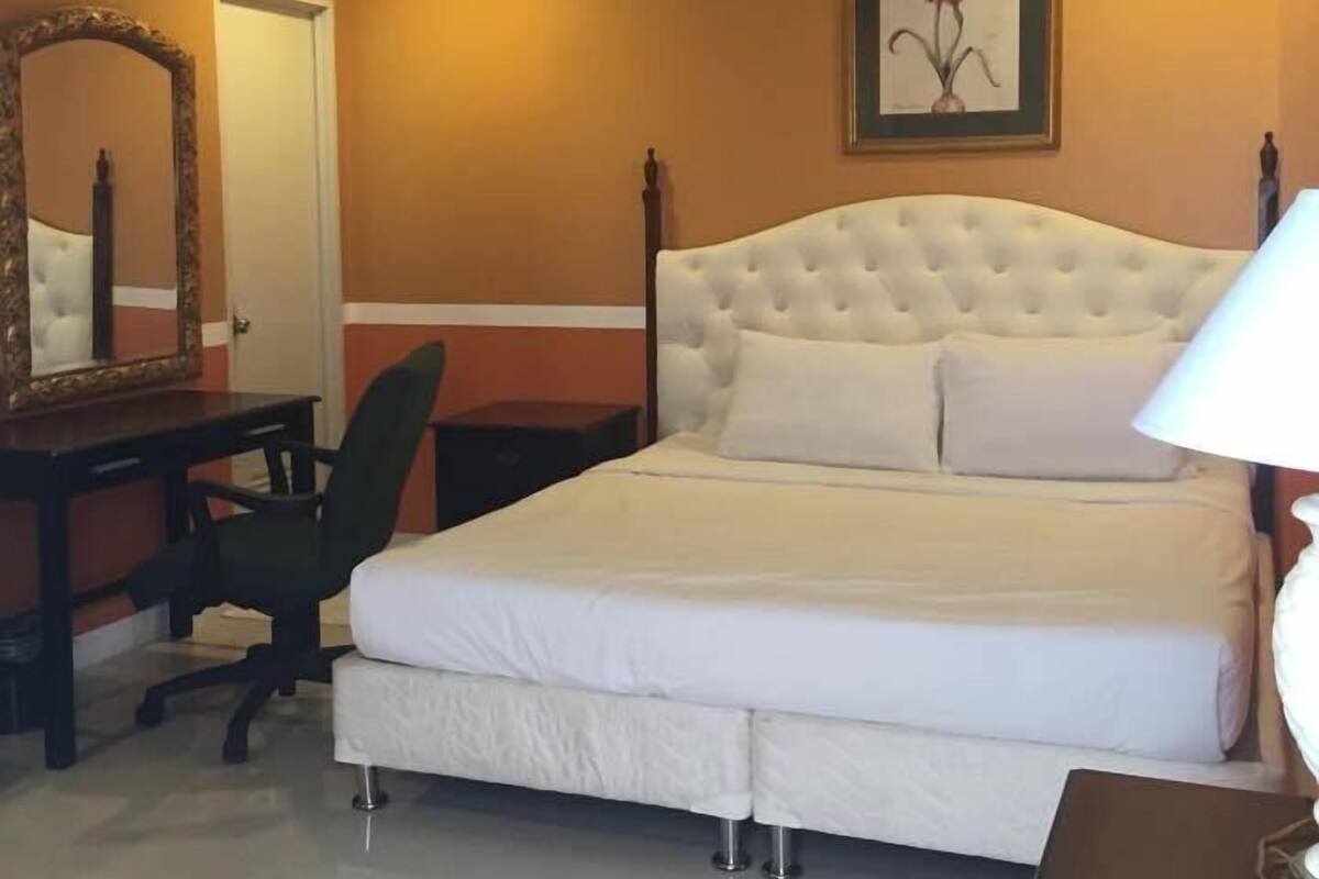 A bedroom with a white bed, a desk with a chair, a mirror, and a framed picture on the wall.