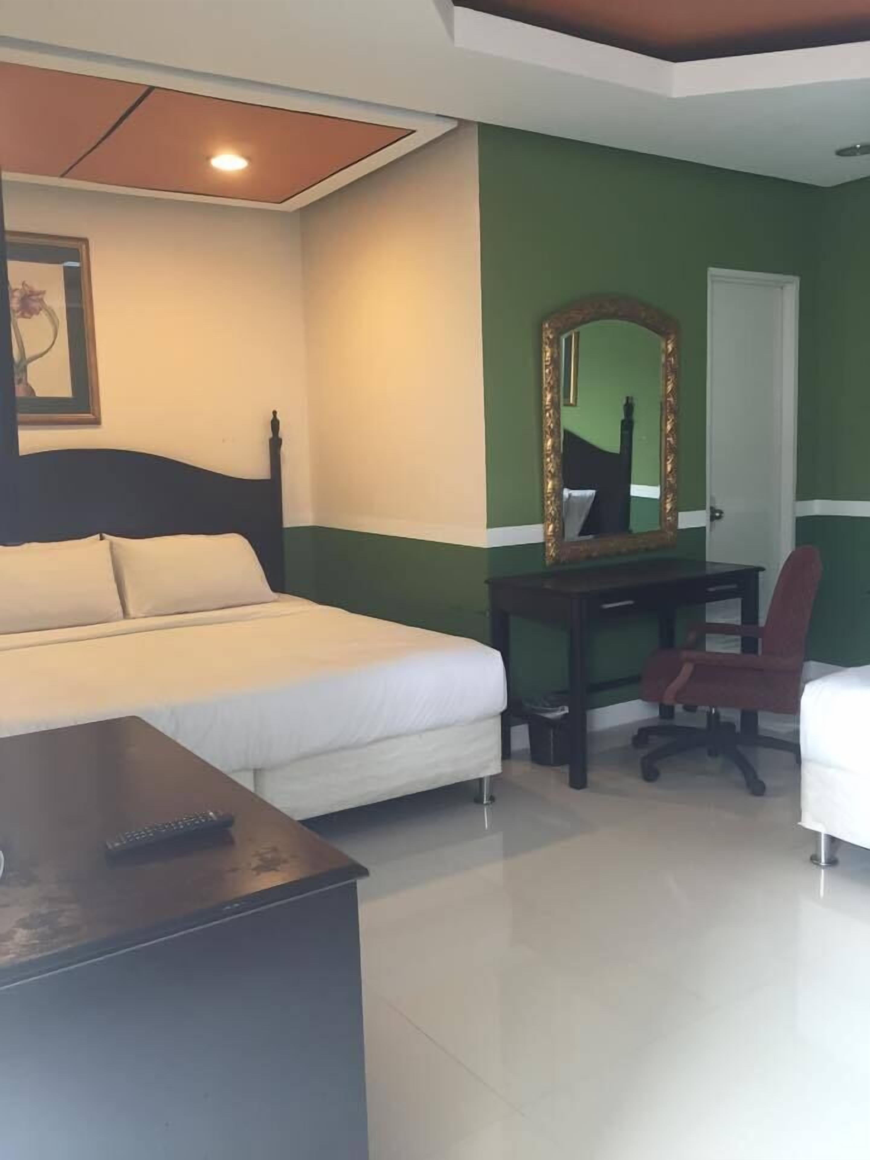 A hotel room with a bed, a desk with a mirror, a chair, and a small table with a remote control.