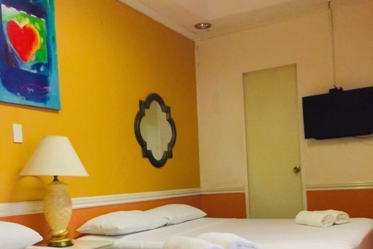A hotel room with two beds, a lamp, a painting, a mirror, and a television on the wall.