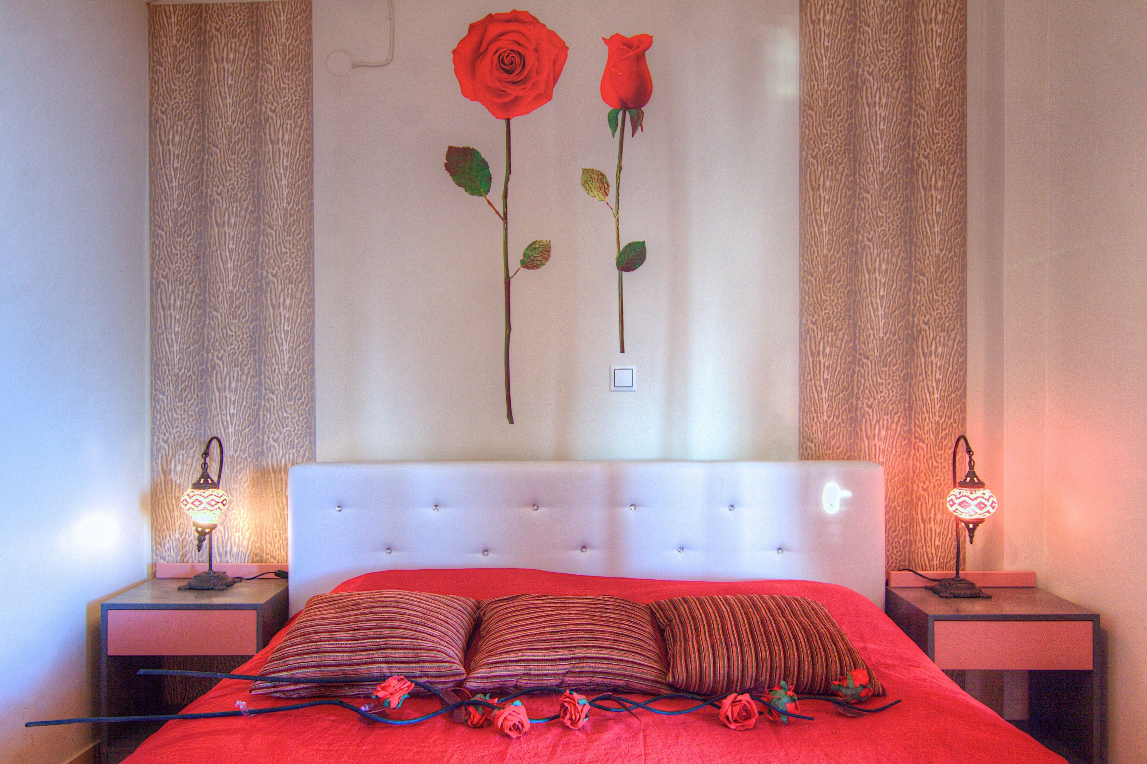 A bedroom with a white tufted headboard, two bedside tables with lamps, a red bedspread, and two artificial red roses on the wall.