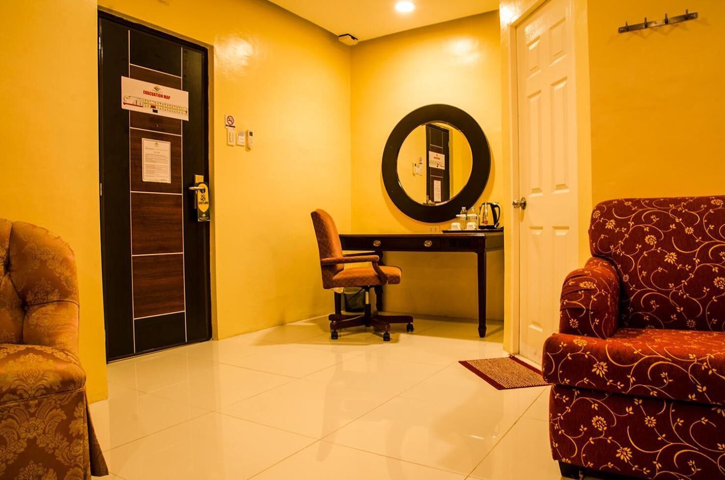 A hotel room with a yellow wall, a desk with a chair, a red patterned armchair, and a door with a sign.
