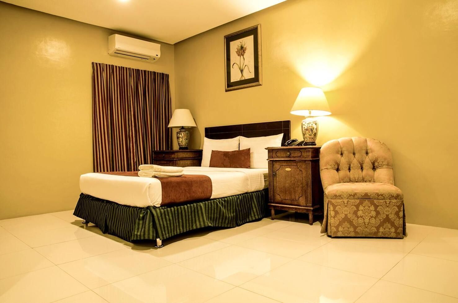 A hotel room with a bed, a nightstand, a chair, a lamp, and an air conditioning unit.