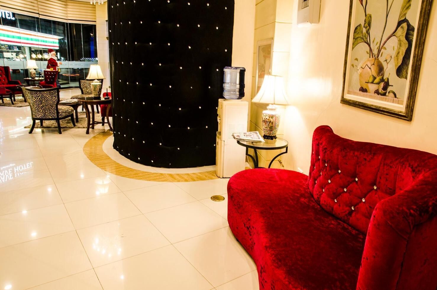 Un vestíbulo de hotel con un sofá de terciopelo rojo, un pilar negro con luces, una mesa con lámpara y un cuadro en la pared.