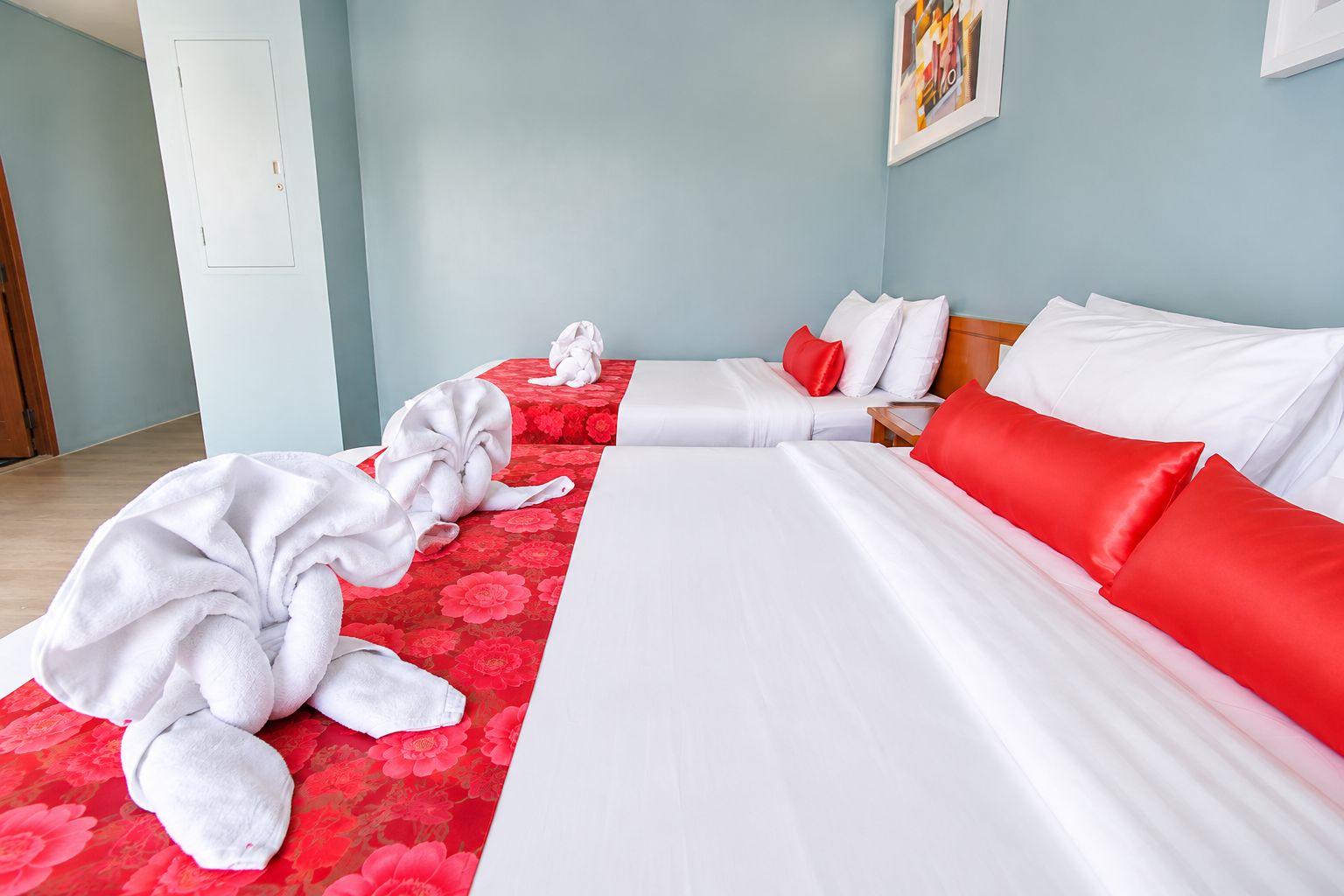 A hotel room with two beds, one with red and white bedding and the other with white bedding. There are towels on the floor and a wooden nightstand with a lamp.